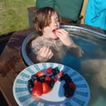 Child relaxing in an outdoor hot tub at a Sussex cabin, eating fresh berries in the sunshine